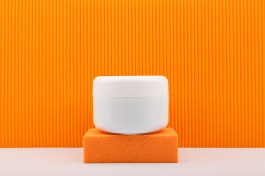 White cosmetic jar on orange podium against orange background. Concept of beauty products with citrus extracts. Cosmetic jar with cream, mask or scrub against colored background with copy space