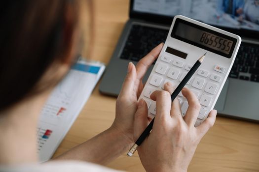 Close up of businesswoman or accountant hand holding pencil working on calculator to calculate financial data report, accountancy document and laptop computer at office, business concept