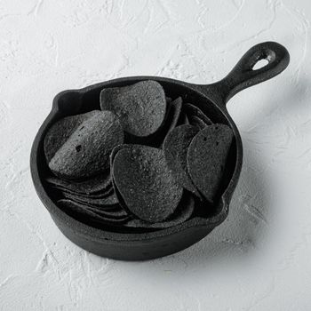 Black crispy potato chips set, in cast iron frying pan, on white stone surface, square format