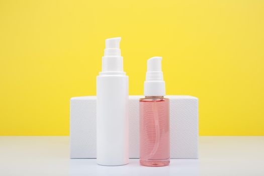 Face cream in white tube and pink cleansing foam in transparent tube against white podium and yellow background with copy space. Concept of daily skin cleaning and moisturizing