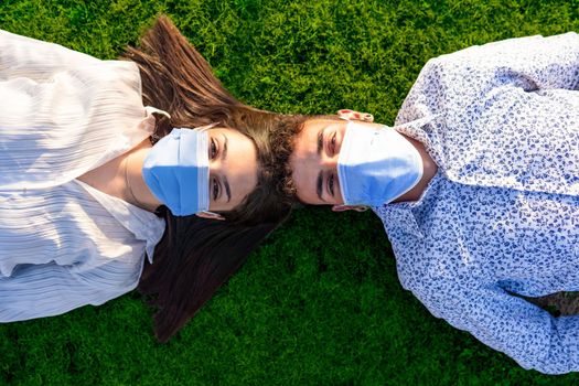 Beautiful couple of young students lying head to head on backs on green lawn of city park looking into the camera wearing the protective mask against the Coronavirus pandemic. New normal health habits