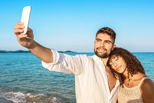 Couple of young lovers taking a selfie on vacation on seashore at sunset or sunrise with boho summer clothes. Afro-American model in love leans her head on the shoulder of her handsome boyfriend
