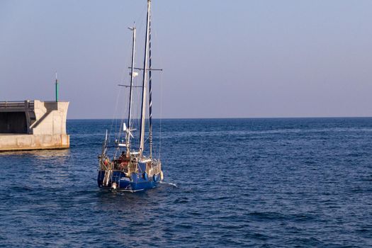 Wide-shot of a blue sailboat leaving port towards open sea on a sunny day at Malaga's Port