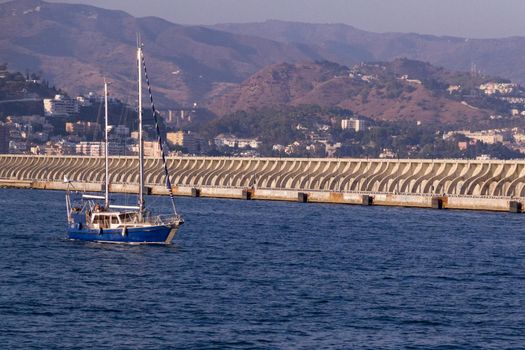 A blue sailboat leaving Malaga's port in front of a dock with the city in the background