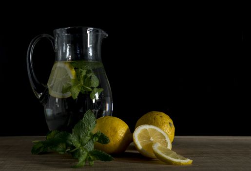 still life with a jug of water filled with mint and lemon with the raw lemon fruit in the foreground on wooden table