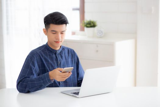 Young asian man working laptop computer and reading smartphone on internet online on desk at home, freelance male smiling using phone with social media on table, business and communication concept.