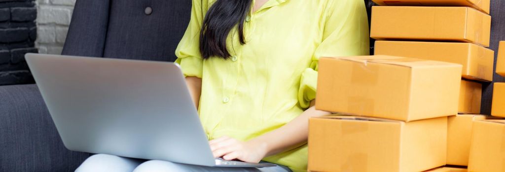 Young asian woman is merchant using laptop computer on sofa checking order of customer with purchase online shopping, business SME or startup, entrepreneur or seller and service delivery product.