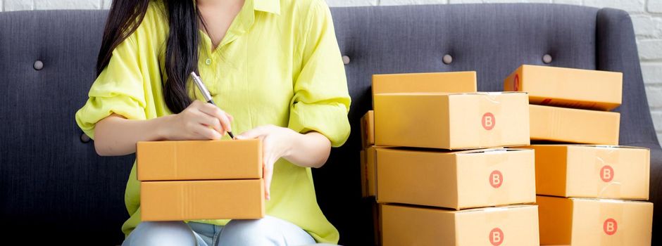 Young asian woman writing address on parcel box at home for delivery order to customer, shipping and logistic, merchant online and seller, business owner or SME, online shopping and e-commerce.