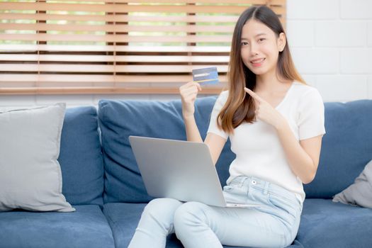 Young asian woman sit on sofa using laptop computer shopping online with credit card buying to internet, happy girl payment with e-business on couch, purchase and payment, business concept.