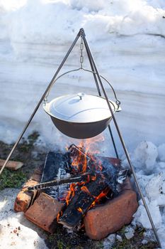 Over the fire hangs a pot in which to cook food. On a hook on a tripod, steam comes out of the pan. Winter Camping outdoor cooking