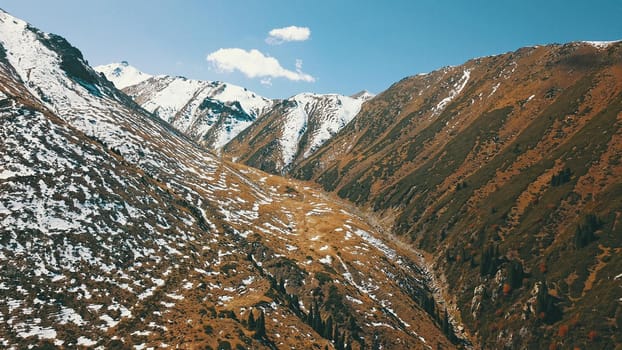 Autumn mountains covered with snow in places. View from the drone, from above. Huge slopes of the gorge, yellow grass and snow. Large stones visible in the river. Shadow of clouds on the ground.