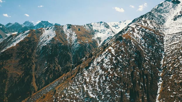 Autumn mountains covered with snow in places. View from the drone, from above. Huge slopes of the gorge, yellow grass and snow. Large stones visible in the river. Shadow of clouds on the ground.