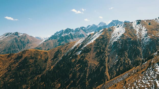 Autumn mountains covered with snow in places. View from the drone, from above. Huge slopes of the gorge, yellow grass and snow. Large stones visible in the river. Shadow of clouds on the ground.