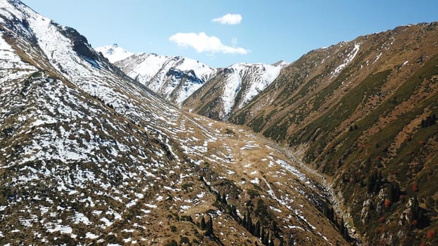 Autumn mountains covered with snow in places. View from the drone, from above. Huge slopes of the gorge, yellow grass and snow. Large stones visible in the river. Shadow of clouds on the ground.