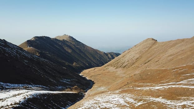 Autumn mountains covered with snow in places. View from the drone, from above. Huge slopes of the gorge, yellow grass and snow. Large stones visible in the river. Shadow of clouds on the ground.