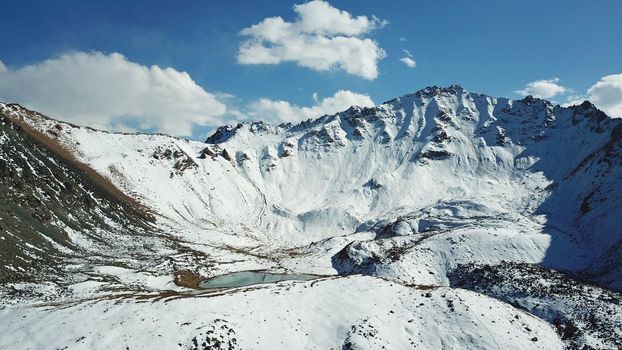 Snow-capped mountains and mountain lake. Top view from a drone. Blue sky and clouds are reflected in the water. Above the mountains. Steep slopes and rocky peaks. The gorge is covered with snow.