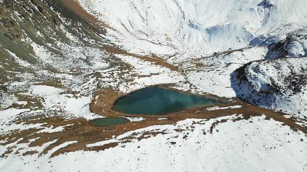 Snow-capped mountains and mountain lake. Top view from a drone. Blue sky and clouds are reflected in the water. Above the mountains. Steep slopes and rocky peaks. The gorge is covered with snow.