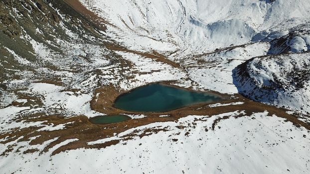 Snow-capped mountains and mountain lake. Top view from a drone. Blue sky and clouds are reflected in the water. Above the mountains. Steep slopes and rocky peaks. The gorge is covered with snow.