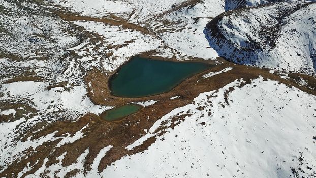 Snow-capped mountains and mountain lake. Top view from a drone. Blue sky and clouds are reflected in the water. Above the mountains. Steep slopes and rocky peaks. The gorge is covered with snow.