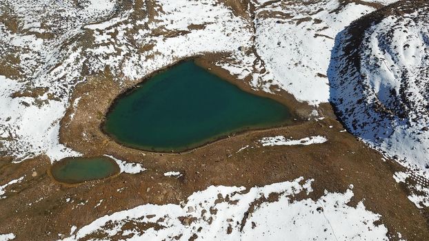 Snow-capped mountains and mountain lake. Top view from a drone. Blue sky and clouds are reflected in the water. Above the mountains. Steep slopes and rocky peaks. The gorge is covered with snow.