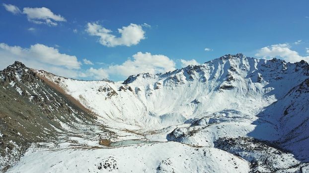 Snow-capped mountains and mountain lake. Top view from a drone. Blue sky and clouds are reflected in the water. Above the mountains. Steep slopes and rocky peaks. The gorge is covered with snow.