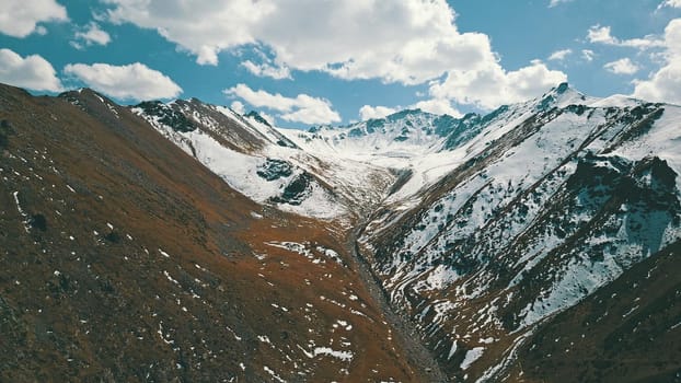 Autumn mountains covered with snow in places. View from the drone, from above. Huge slopes of the gorge, yellow grass and snow. Large stones visible in the river. Shadow of clouds on the ground.