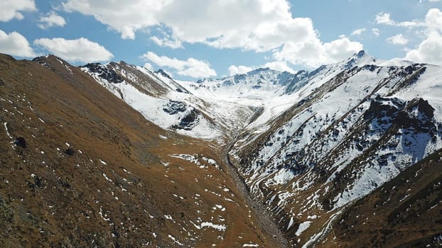 Autumn mountains covered with snow in places. View from the drone, from above. Huge slopes of the gorge, yellow grass and snow. Large stones visible in the river. Shadow of clouds on the ground.