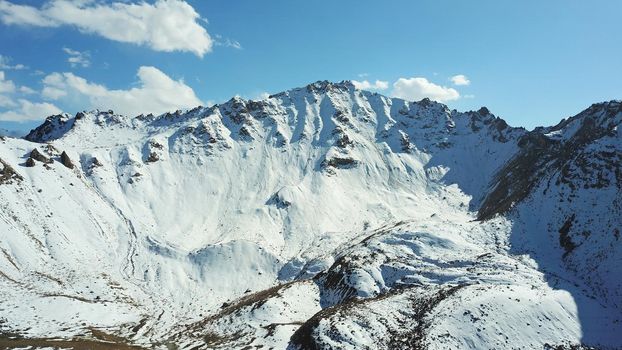 Snow-capped mountains and mountain lake. Top view from a drone. Blue sky and clouds are reflected in the water. Above the mountains. Steep slopes and rocky peaks. The gorge is covered with snow.