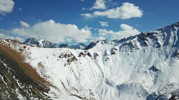 Snow-capped mountains and mountain lake. Top view from a drone. Blue sky and clouds are reflected in the water. Above the mountains. Steep slopes and rocky peaks. The gorge is covered with snow.