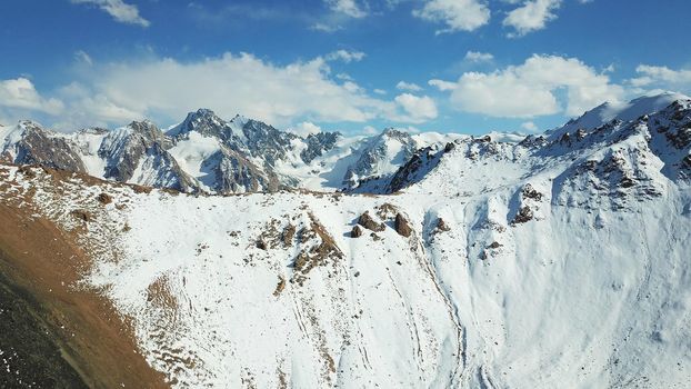 Snow-capped mountains and mountain lake. Top view from a drone. Blue sky and clouds are reflected in the water. Above the mountains. Steep slopes and rocky peaks. The gorge is covered with snow.