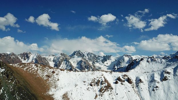 Snow-capped mountains and mountain lake. Top view from a drone. Blue sky and clouds are reflected in the water. Above the mountains. Steep slopes and rocky peaks. The gorge is covered with snow.