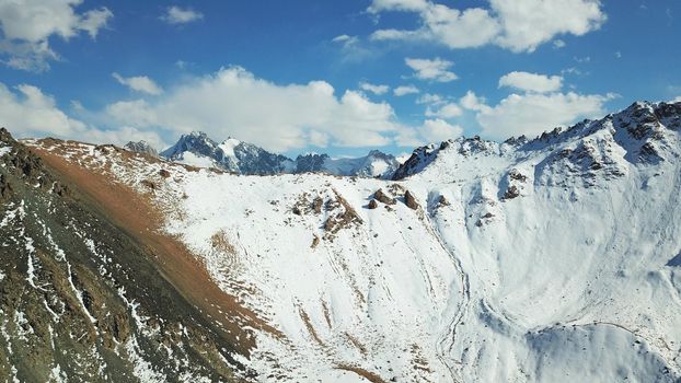 Snow-capped mountains and mountain lake. Top view from a drone. Blue sky and clouds are reflected in the water. Above the mountains. Steep slopes and rocky peaks. The gorge is covered with snow.