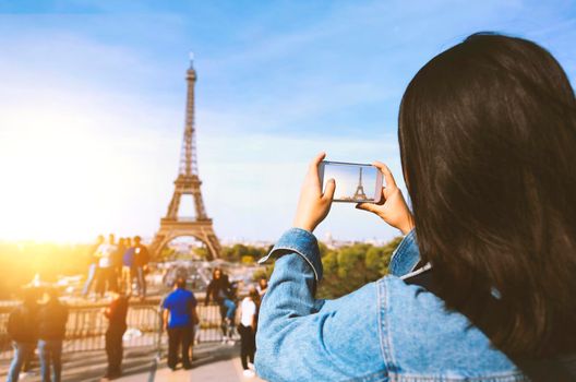 Woman tourist taking photo by phone near the Eiffel tower in Paris under sunlight and blue sky. Famous popular touristic place in the world