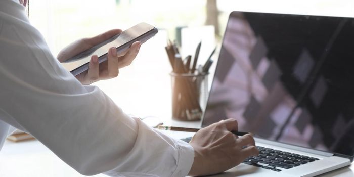 Close up of hand young female using mobile phone to searching information while she working with laptop on desk.