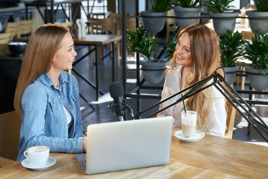 Side view of two young attractive blogger women sitting in cafe and communicating online with followers by laptop. Concept of talking about different topics with modern microphone. 