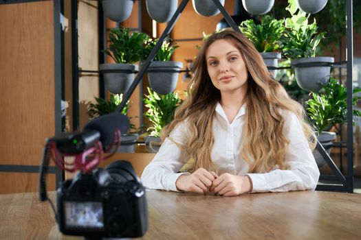 Front view of young beautiful woman blogger with long hair sitting at the table in cafe and preparing for interview on camera. Concept of process interview in modern cafe. 