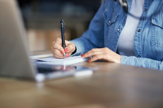 Close up of woman in denim jacket holding pen and writing important information in notebook. Concept of process preparing for communication about interesting facts. 