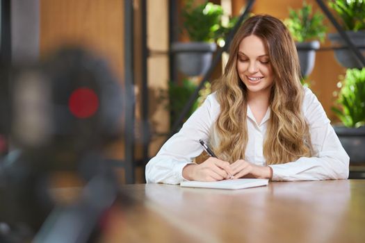 Front view portrait of smiling pretty young woman with long hair in white shirt sitting ar the table and working with notebook and pen. Concept of process working with good mood in cafe. 