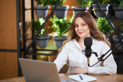 Front view of attractive young blogger woman in white shirt sitting at the table in cafe with notebook, modern microphone and laptop. Concept of process preparing for interview online. 