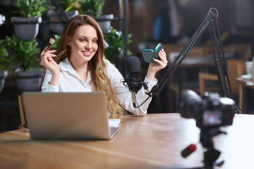 Front view of smiling young woman in white shirt doing makeup in front of the camera in cafe. Concept of process preparing for communicating online with followers by laptop. 