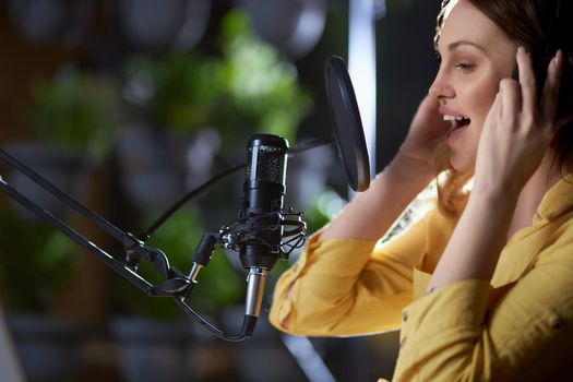 Side view portrait of attractive young woman in modern headphone singing into black microphone. Concept of process recording trendy songs in professional music studio. 