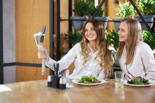 Side view of happy young attractive blogger women eating tasty salads in cafe and communicating with friends online on phone. Concept of free time with delicious food and with good mood. 