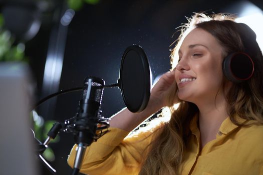 Close up portrait of young beautiful woman in yellow shirt singing with headphone into black modern microphone. Concept of process recording song in professional studio. 