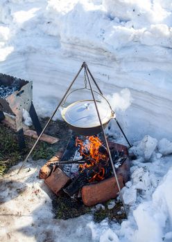 Over the fire hangs a pot in which to cook food. On a hook on a tripod, steam comes out of the pan. Winter Camping outdoor cooking