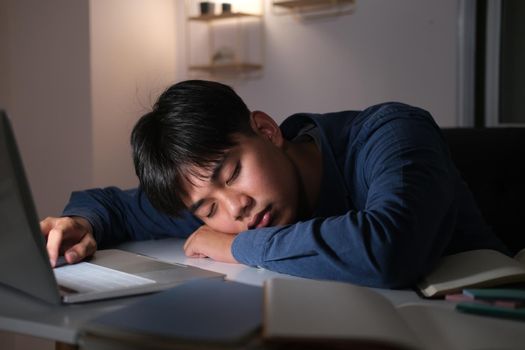 Sleepy exhausted collage young man working overtime at office desk with his laptop, he is about to fall asleep, sleep deprivation and overtime working concept.