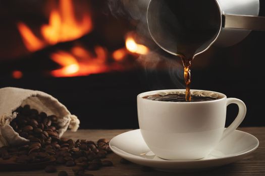 Pouring cezve coffee into a cup on a wooden table in front of a burning fireplace. Selective focus.