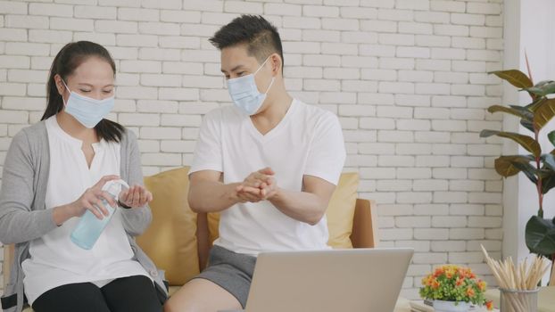 Happy Asian beautiful family couple husband and wife laughing sitting on sofa in the living room working with laptop computer at home. Woman brings alcohol gel applying to hand man, COVID-19