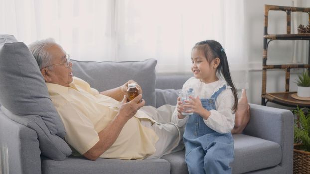 Asian granddaughter brought medicine and water for Grandpa to eat on the sofa in the lounge, Senior old man taking pills medicine time, granddaughter take care older with love