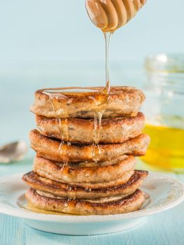 Stack of small pancakes with banana and chia seeds in honey on light blue background. Honey pouring over stack of mini pancakes with chia or poppy seeds.