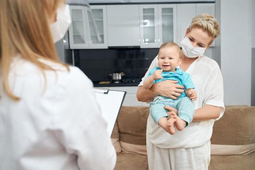 Female doctor writing something on clipboard and mother standing near and holding baby on hands. Pediatrician in protective mask visiting little patient at home.